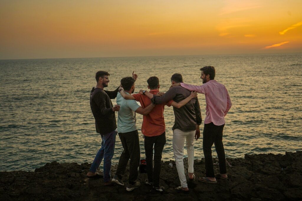 Five men with arms around each other on a rocky shore at golden hour